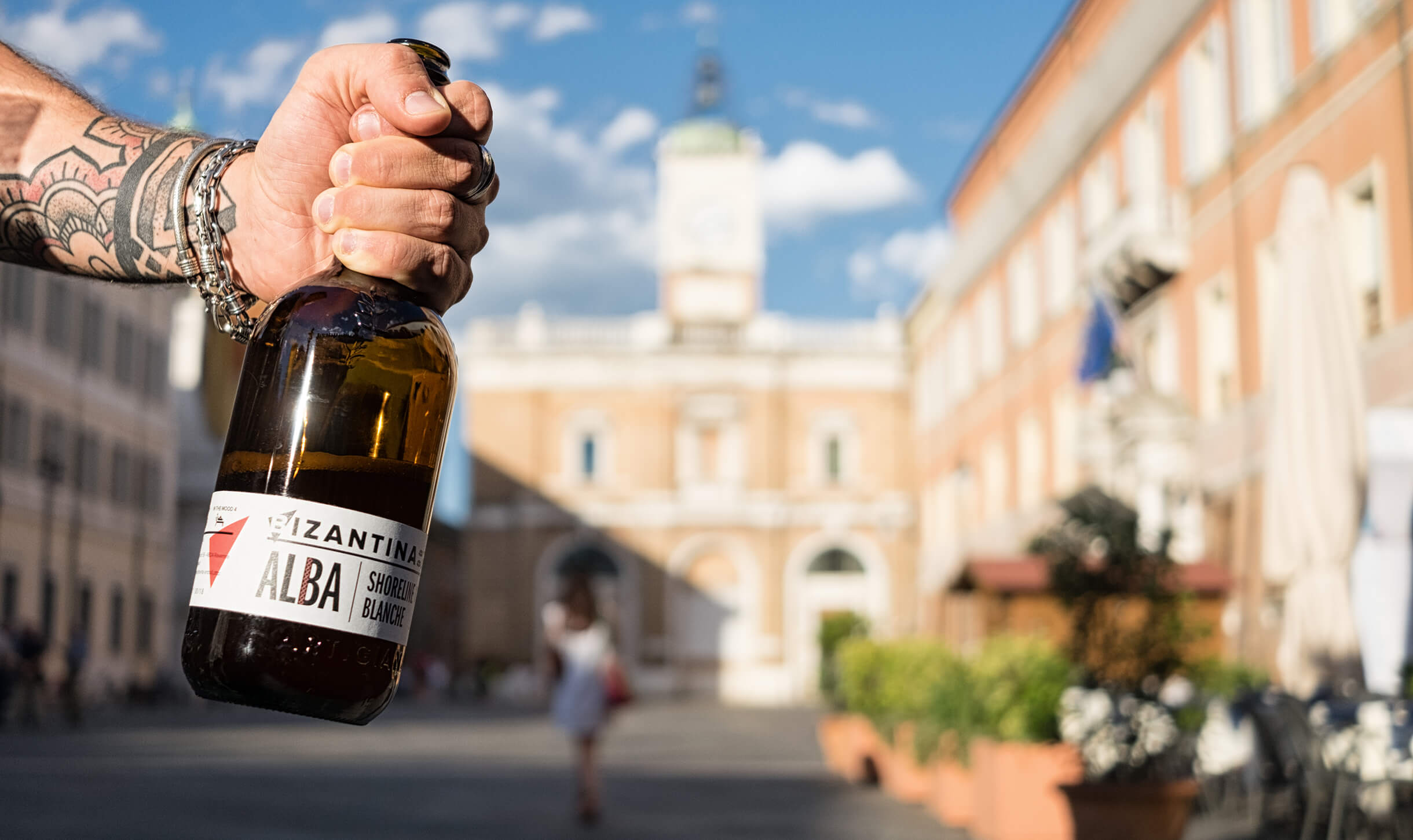 primo piano di una mano che tiene una birra sullo sfondo di Piazza del Popolo a Ravenna