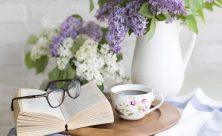 Libro aperto con occhiali appoggiati vicino ad una tazzina da caffè e sullo sfondo dei fiori